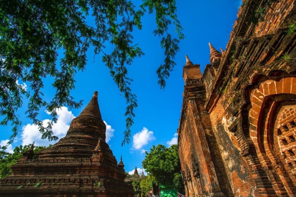 Is Bagan a Spiritual Place? Discover Why Should You Visit Bagan Myanmar in 2026! This image captures the historical essence of Bagan, Myanmar, showing a close-up view of aged, reddish-brown brick pagodas under a vibrant blue sky. Overhanging branches with green leaves frame the top of the image, adding a natural, serene touch to the scene of these ancient Buddhist structures.