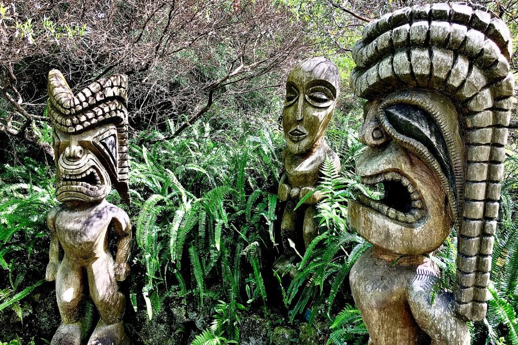 Discovering the Spiritual Essence of Haleakalā Volcano Hawaii Three intricate wooden tiki statues stand amidst lush ferns, with expressions that are a blend of fierce and serene, capturing the essence of Polynesian cultural art. They are set against a backdrop of dense tropical foliage, highlighting the art's deep connection to nature and the rich heritage of the Hawaiian islands.