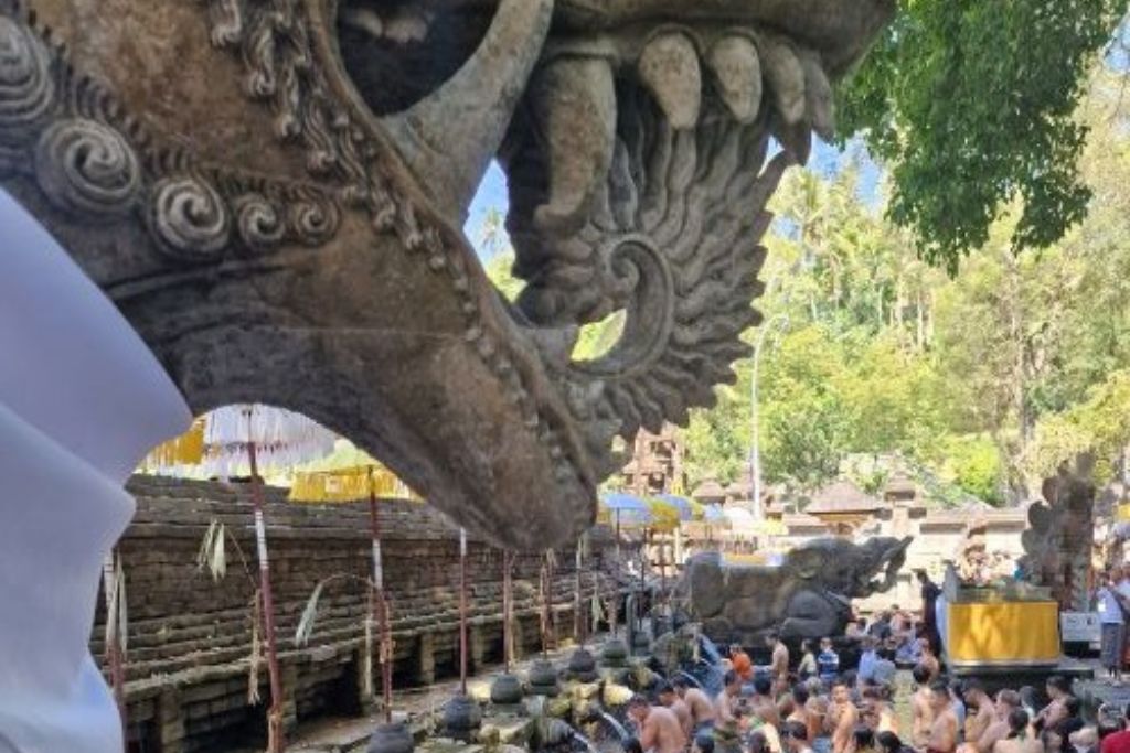 This image shows a traditional ceremony at a temple in Bali, Indonesia, viewed through a beautifully detailed stone carving. In the foreground, the intricate sculpture frames a bustling scene of numerous attendees in traditional attire, engaged in a religious ritual. The background features tropical greenery and more stone temple architecture, enhancing the exotic and spiritual atmosphere of the event.