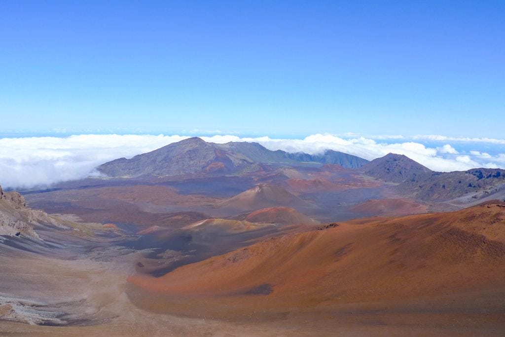 Discovering the Spiritual Essence of Haleakalā Volcano Hawaii A sweeping view of the Haleakalā Crater in Maui, Hawaii, showcasing the vast, colorful volcanic landscape with hues of red, brown, and gray. The crater's rugged terrain is surrounded by clouds at the elevation where this photo was taken, giving a sense of the grand scale and isolated beauty of this ancient volcanic caldera.