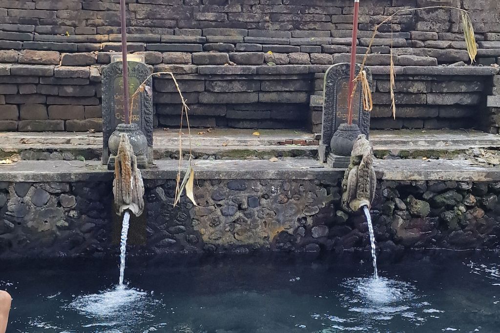 Tirta Empul Temple, the Sacred Water Temple in Ubud (Healing Guide) The image captures a serene scene at Tirta Empul Temple in Bali, Indonesia, featuring a sacred water pool. Two water spouts crafted from stone pour crystal clear water into the pool, while traditional Balinese umbrellas in golden yellow and white with intricate lace detail stand gracefully in the background. The stone steps leading to the pool, along with the lush greenery partially visible, add to the tranquility and cultural richness of the setting.