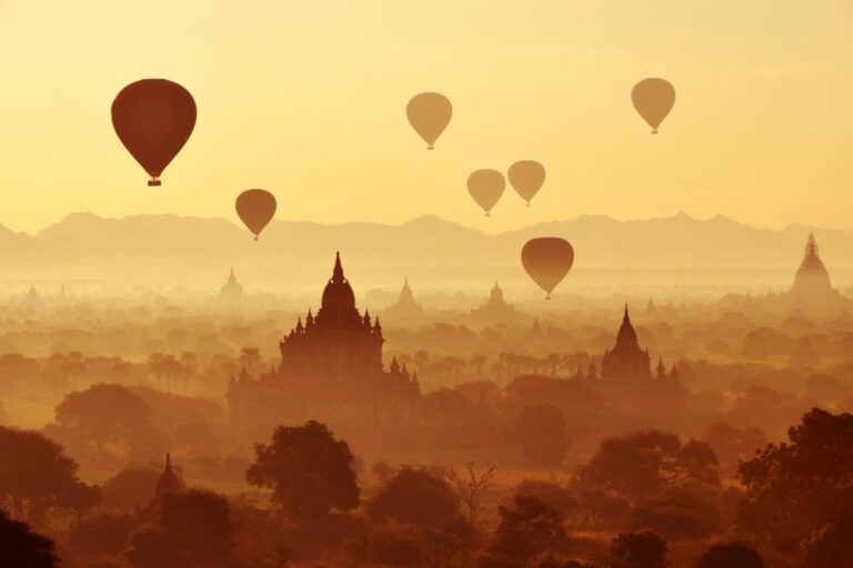 This image depicts multiple hot air balloons floating above a misty, historic landscape of Bagan at sunrise. The scene captures a serene view of ancient temples and scattered trees silhouetted against a golden sky, with distant mountains providing a picturesque backdrop.