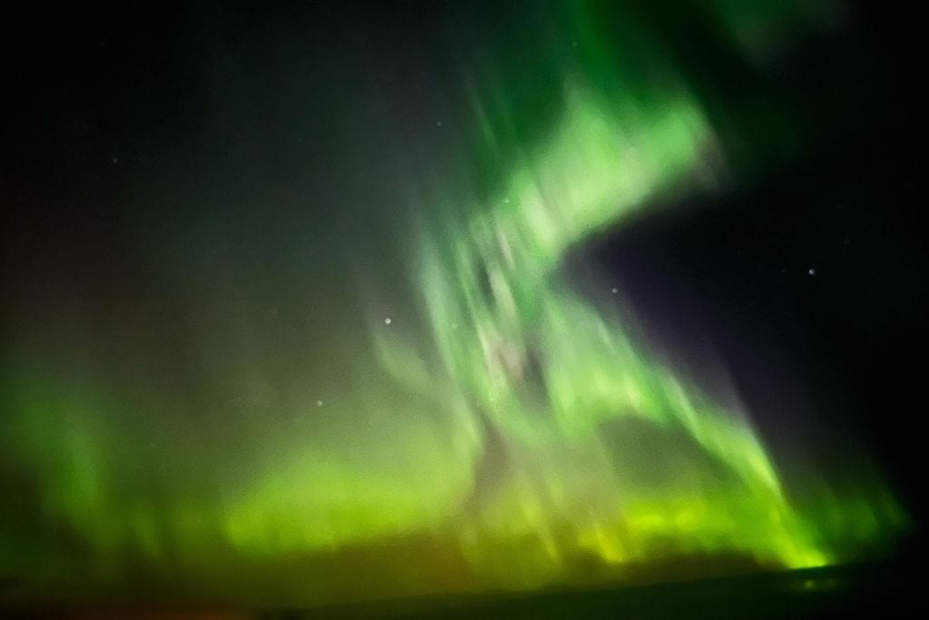 How to Plan a Trip to See Aurora Borealis? My Personal Experience on Spotting Magical Northern Lights in 2026! This image captures the vivid and dynamic display of the aurora borealis. Brilliant green lights swirl through the night sky, creating ethereal shapes above a barely visible horizon. The scene is set against a backdrop of a dark, starlit sky, enhancing the luminous and otherworldly appearance of the northern lights.