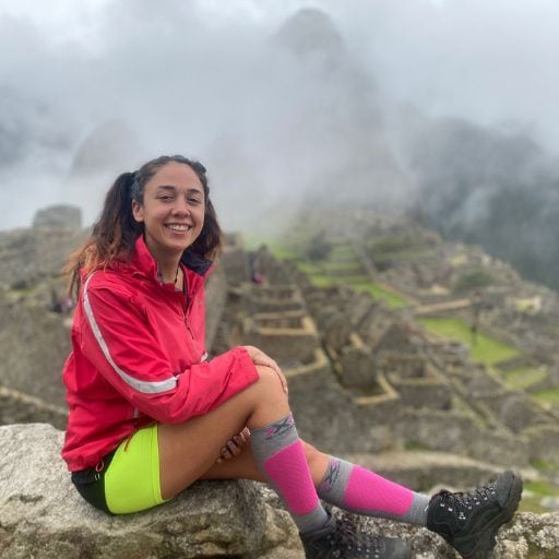 A young woman with a bright smile sitting on a rock at Machu Picchu, wearing a pink rain jacket, neon green shorts, and pink socks. Misty mountains and the ancient Incan ruins are visible in the background.