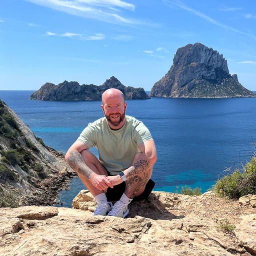 Tom Henty, the author man squatting on a rocky outcrop, smiling at the camera, with the calm blue sea and a towering rocky island in the background under a clear blue sky. He's wearing a light green t-shirt, shorts, and sneakers, with a tattoo visible on his left arm.