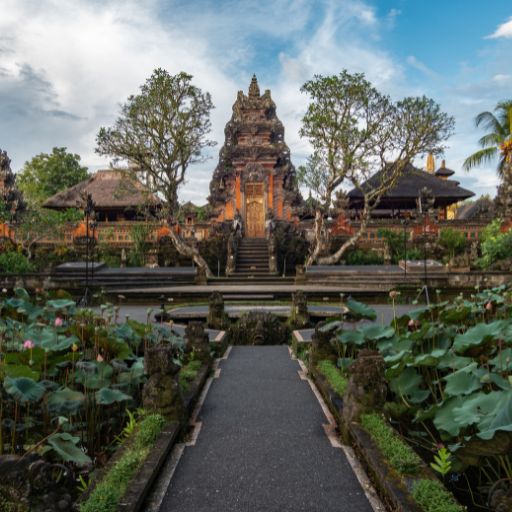 Tirta Empul Temple, the Sacred Water Temple in Ubud (Healing Guide) This image features a scenic view of a traditional Balinese temple gate framed by lush greenery and a pond with lotus plants. The path leading up to the ornate, towering gate is lined by stone statues, emphasizing the cultural and architectural heritage of Bali.