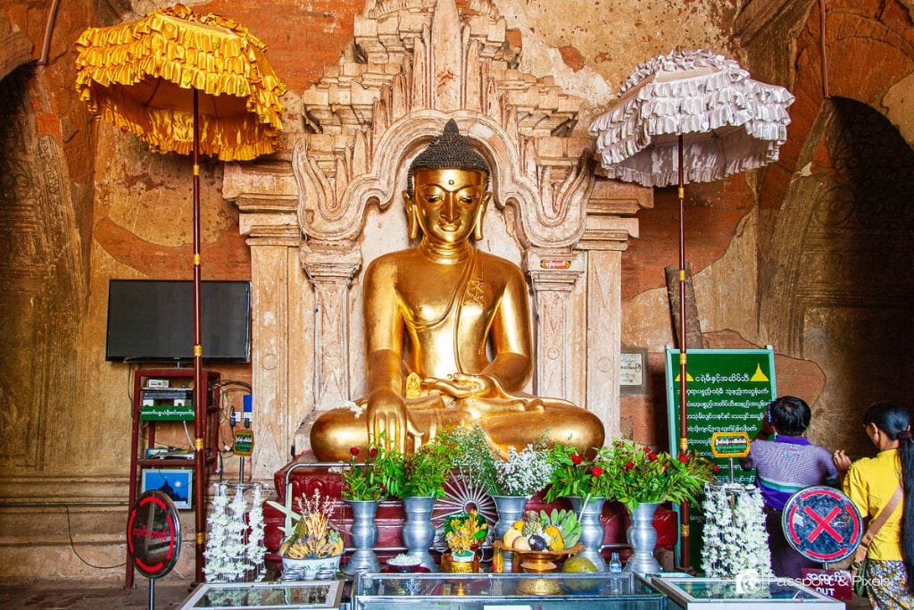 Is Bagan a Spiritual Place? Discover Why Should You Visit Bagan Myanmar in 2026! Inside a temple in Bagan, Myanmar, showing a large golden Buddha statue adorned with flowers and ceremonial umbrellas, with devotees praying in front.
