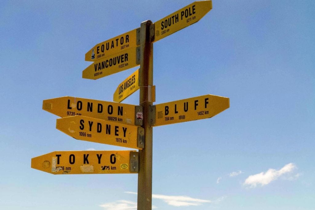 Signpost at Cape Reinga displaying distances to various global cities such as London, Sydney, and Tokyo, under a clear blue sky.
