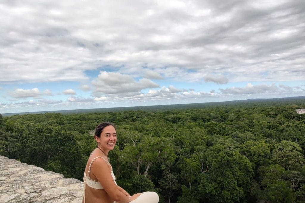 The image features a smiling woman sitting atop an ancient structure, enjoying a panoramic view of an expansive rainforest that stretches into the horizon under a cloudy sky. Her casual pose and joyful expression convey a sense of adventure and appreciation for the breathtaking natural surroundings, offering a personal touch to the stunning landscape. This setting likely suggests a visit to the historical site of Tikal in a tropical location, emphasizing the vastness and beauty of the natural world.