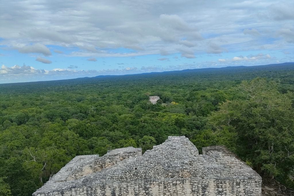 The image offers a dramatic aerial view of an ancient Mayan ruin, showcasing the intricate stone architecture and multiple levels of the temple against a vast backdrop of dense rainforest. Several people can be observed exploring the site, which emphasizes the scale and historical significance of the location. Overhead, a dramatic sky filled with thick clouds adds a dynamic element to the scene, further enhancing the sense of discovery and majesty of this cultural landmark.