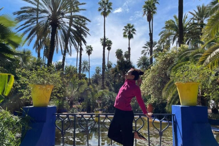 A woman in a pink jacket and black pants is performing a yoga pose on a blue-railed bridge in a lush garden filled with tall palm trees and vibrant green foliage. Two large yellow pots with plants flank the bridge, and a serene pond lies in the background under a bright blue sky.