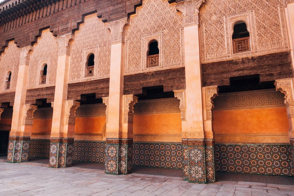 The Seven Saints of Marrakech (Local Insight of Marrakesh) A beautiful facade of a building adorned with intricate carvings and geometric mosaic tiles. The arched windows and doorways are framed by detailed patterns, showcasing traditional architectural craftsmanship. The warm hues of the walls and the elaborate tilework create a striking visual contrast.