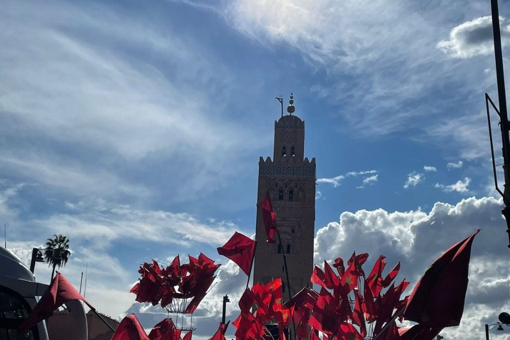 The Seven Saints of Marrakech (Local Insight of Marrakesh) A tall minaret with ornate details rises against a backdrop of a bright blue sky with scattered clouds. In the foreground, numerous red flags flutter, adding vibrant color and movement to the scene. The composition captures a striking contrast between the traditional architecture and the dynamic display of flags.