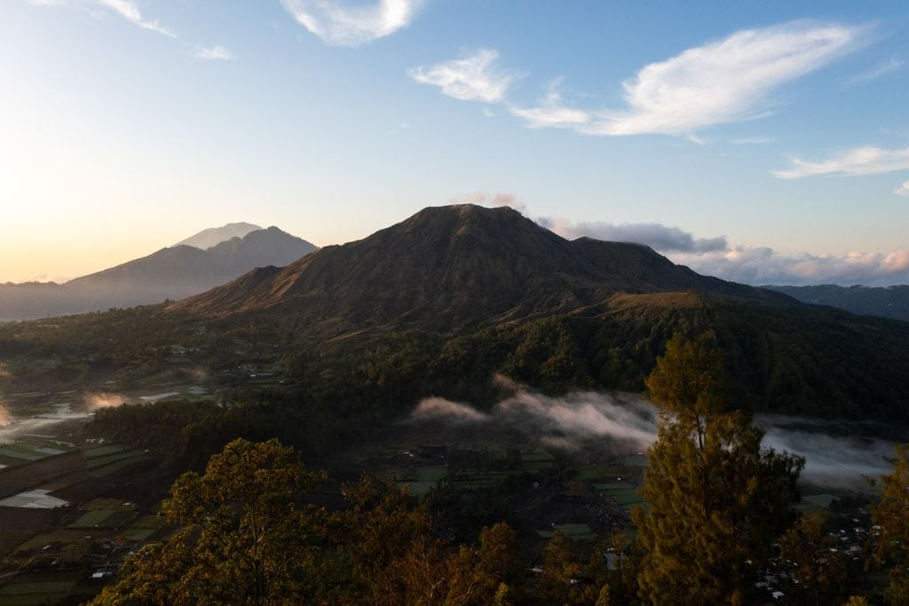 A breathtaking sunrise over Mount Batur, with Mount Abung and Mount Agung visible in the background. The scene captures the early morning light illuminating the peaks and the valley below, with mist gently rising from the forests and fields.