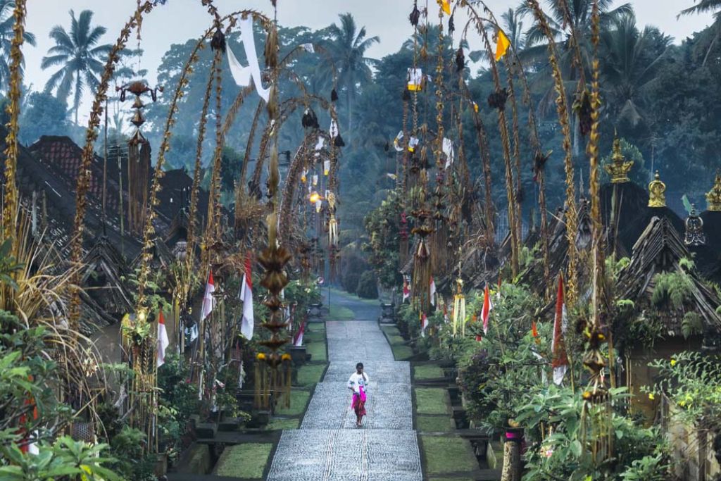 Mount Batur Sunrise Trekking: A Spiritual Journey A Balinese woman walks down a decorated pathway in Penglipuran Village, flanked by tall, arched bamboo poles adorned with traditional decorations. The village is lush with greenery and palm trees, creating a serene and picturesque atmosphere.