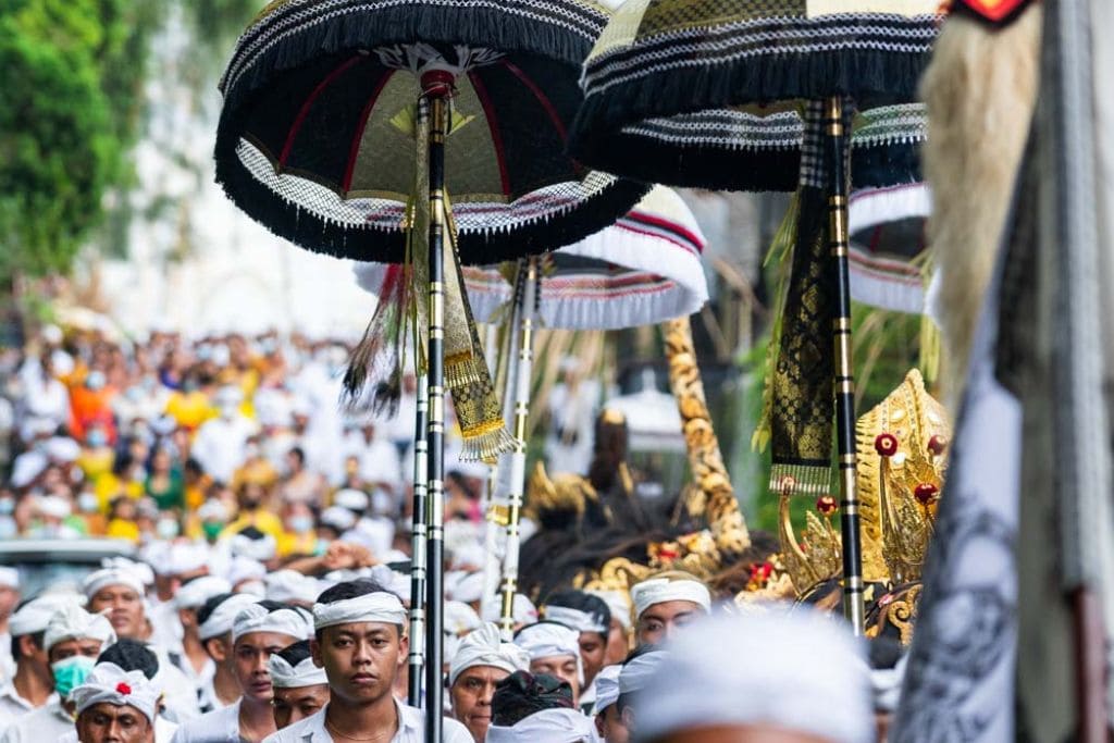 Mount Batur Sunrise Trekking: A Spiritual Journey A vibrant parade during a traditional ceremony in Ubud, Bali, featuring participants dressed in white and carrying ornate black-and-gold umbrellas. The procession is filled with people walking closely together, showcasing cultural attire and ceremonial items.