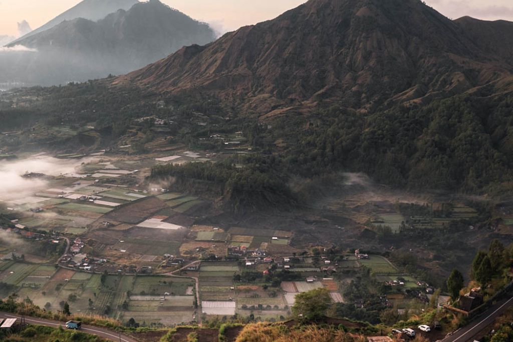 Mount Batur Sunrise Trekking: A Spiritual Journey A scenic view of farmland surrounding Mount Batur in Kintamani, with terraced fields and small buildings dotting the landscape. The winding road in the foreground leads towards the towering mountain, under a sky with soft clouds, capturing the serene and picturesque rural setting.