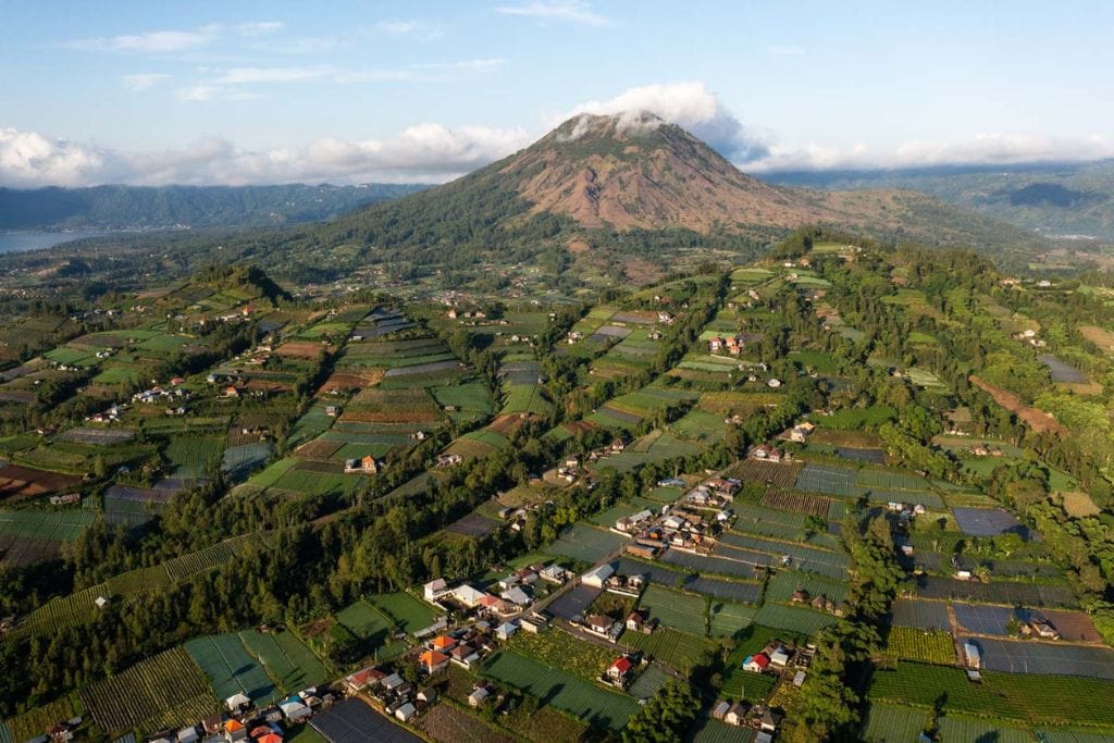 Mount Batur Sunrise Trekking: A Spiritual Journey An aerial view of the lush agricultural landscape surrounding Mount Batur in Kintamani. The patchwork of fields, farms, and small villages stretches out towards the mountain, which stands majestically under a clear sky with a few clouds. The scene captures the harmony between the natural environment and human habitation.