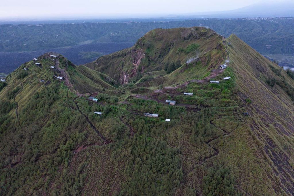 Mount Batur Sunrise Trekking: A Spiritual Journey An aerial view of the summit of Mount Batur at sunrise, showcasing a rugged landscape with small shelters dotting the slopes. The paths winding through the greenery lead to various lookout points, offering panoramic views of the surrounding valleys and distant mountains. The early morning light adds a serene and ethereal quality to the scene.