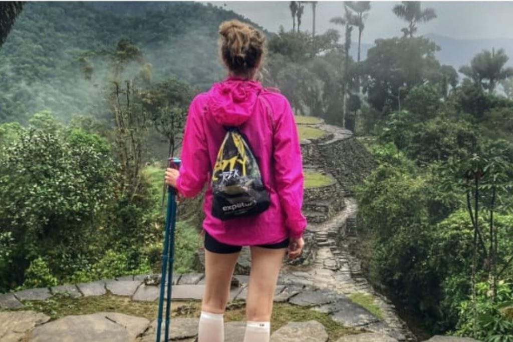 How to Get to the Lost City Colombia? My Personal Experience in 2026! A hiker dressed in a pink jacket and shorts, standing on the ancient stone terraces of Ciudad Perdida, holding a trekking pole and looking out over the lush, mist-covered jungle and mountains of the Sierra Nevada de Santa Marta.