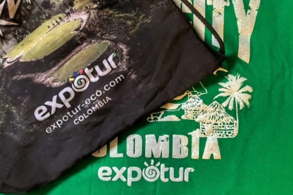 How to Get to the Lost City Colombia? My Personal Experience in 2026! Merchandise from Expotur, including a black drawstring bag and a green T-shirt with the Expotur logo and the word "Colombia," displayed on a flat surface.