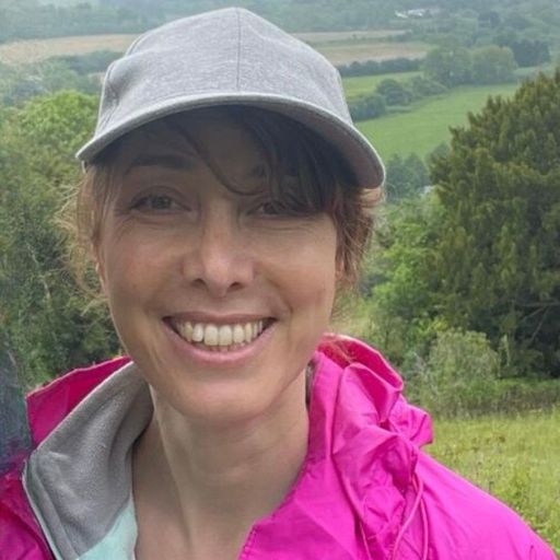 How to Get to the Lost City Colombia? My Personal Experience in 2026! The smiling author of this blog post wearing a gray cap and a pink jacket, standing outdoors with a scenic background of lush green fields and trees. The person appears happy and relaxed, enjoying a day out in nature.