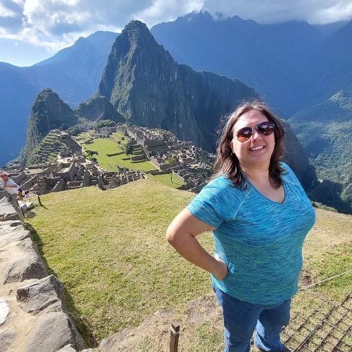 Brodi Cole, a woman with sunglasses stands smiling in the foreground, her arms akimbo, as she poses at Machu Picchu. The ancient Incan ruins stretch out behind her, set against a dramatic backdrop of steep, lush green mountains under a bright blue sky. This image captures a joyful moment of exploration amidst one of the world's most iconic archaeological sites.