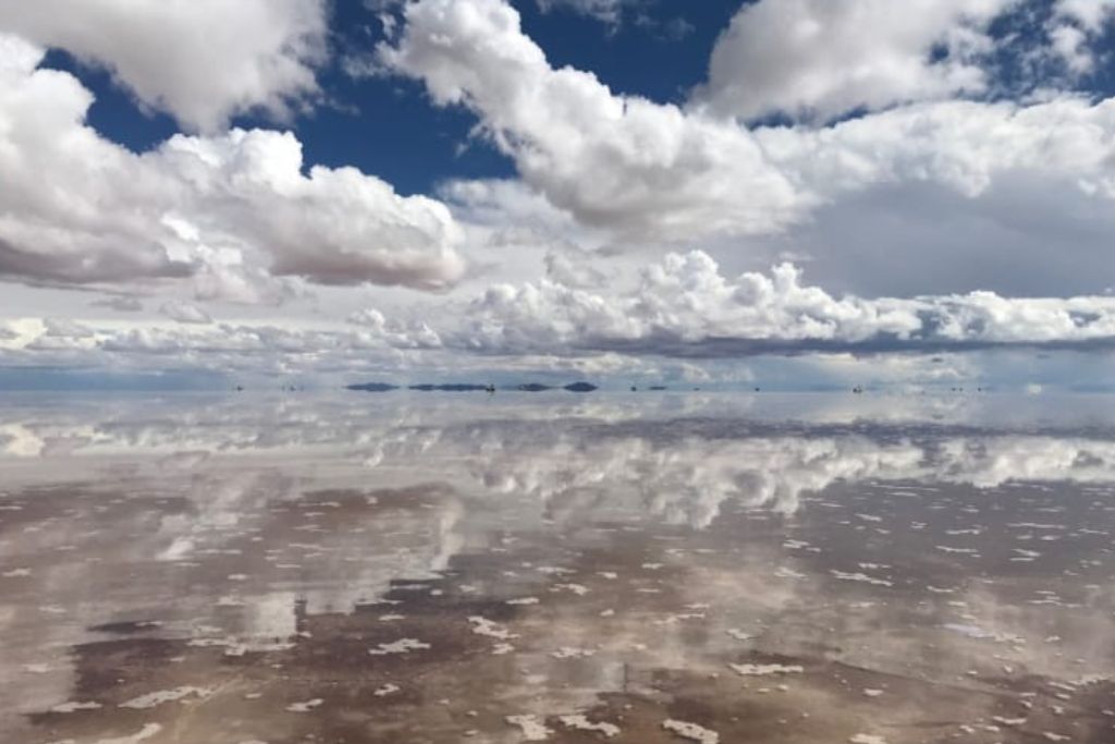 How to Visit Salar de Uyuni in 2026: My Lifetime Experience! An expansive salt flat reflects the cloudy sky above, creating a stunning mirror effect that blends the earth and sky seamlessly. The white clouds, varying in size and density, add depth and texture to the scene, while the distant mountains provide a faint outline on the horizon. The reflective surface creates an ethereal, almost otherworldly landscape.