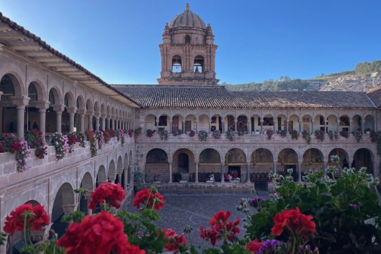A historic courtyard with arches, adorned with vibrant red and pink flowers, leads to a central bell tower under a clear blue sky. The setting suggests a tranquil and picturesque location, the historic site of Cusco mysticism.
