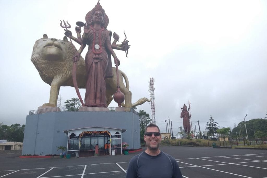 Grand Bassin Sacred Lake: A Sanctuary of Peace & Serenity A man smiling in front of the impressive statues at Grand Bassin Sacred Lake in Mauritius. The large statues include a prominent figure of the Hindu goddess Durga, standing on a lion, and another tall statue of Lord Shiva holding a trident in the background.