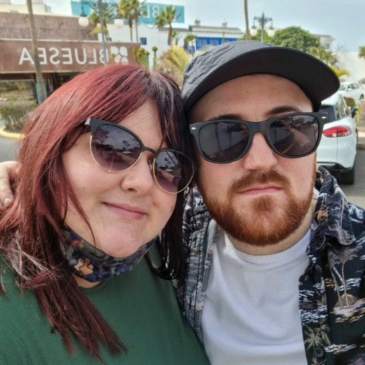 Legends Es Vedra's Spiritual Significance (Ibiza's Mystery) A couple posing for a selfie outside on a sunny day. Both are wearing sunglasses; the woman has red hair and is wearing a green top, while the man has a beard, a black cap, and a patterned shirt. In the background, there's a sign that reads "BLUESA."