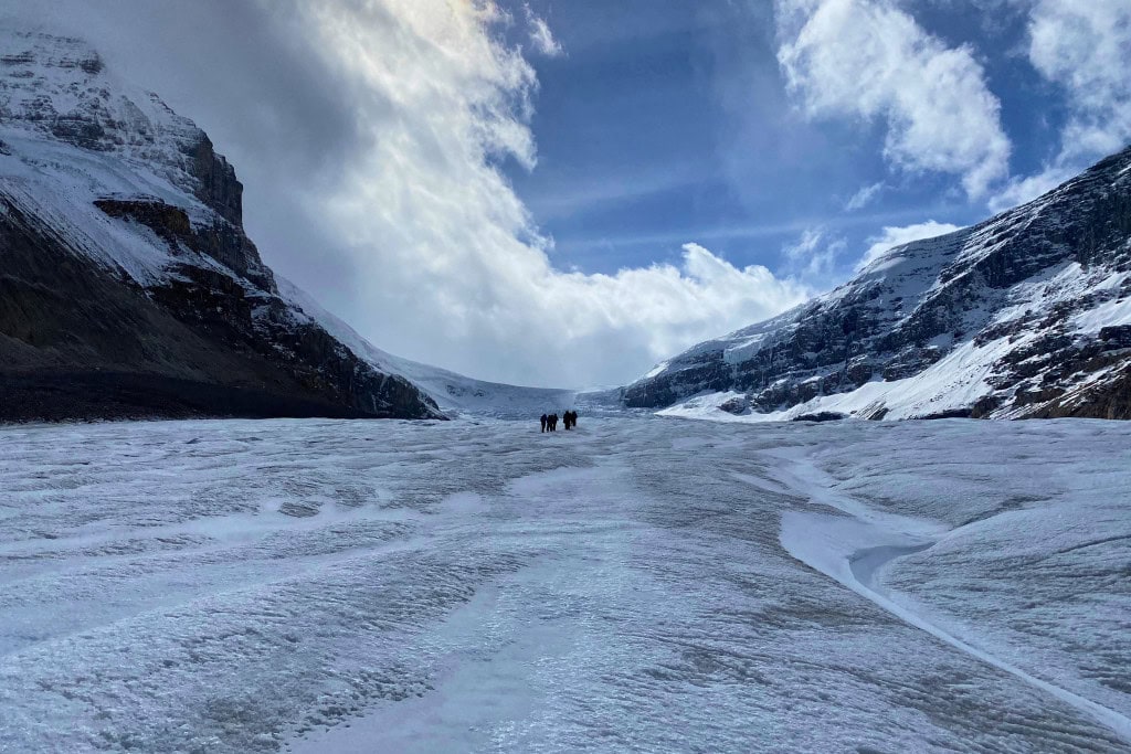 Is Banff Worth Visiting? Explore This Spiritual Place in 2026 A group of hikers walking across the vast, icy expanse of the Athabasca Glacier along the Icefields Parkway. The glacier is surrounded by steep, snow-covered mountains under a partly cloudy sky, highlighting the dramatic and rugged beauty of the area.