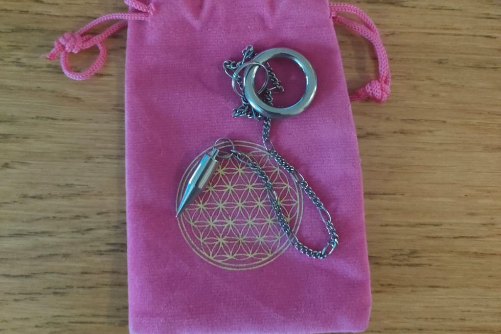 A silver pendulum with a conical-shaped weight and chain, placed on a pink drawstring pouch. The pouch has a geometric flower-like design printed in gold on the front, resting on a wooden surface.