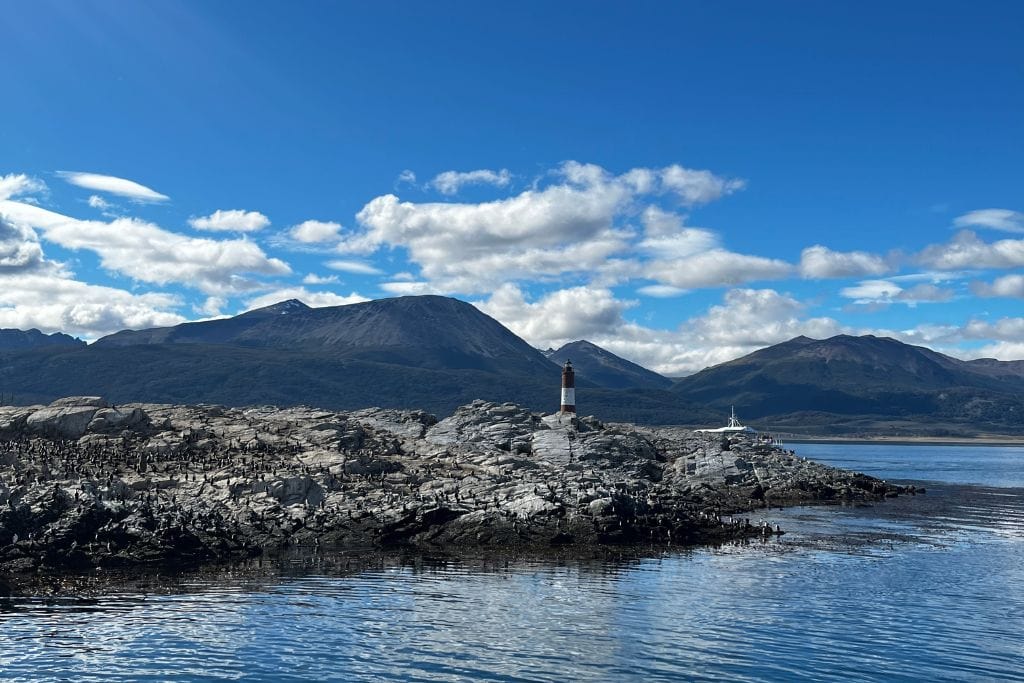 A solitary lighthouse stands on a rocky outcrop surrounded by calm, deep blue water under a clear sky. In the background, majestic mountains rise against a backdrop of scattered fluffy clouds, creating a peaceful and remote landscape.