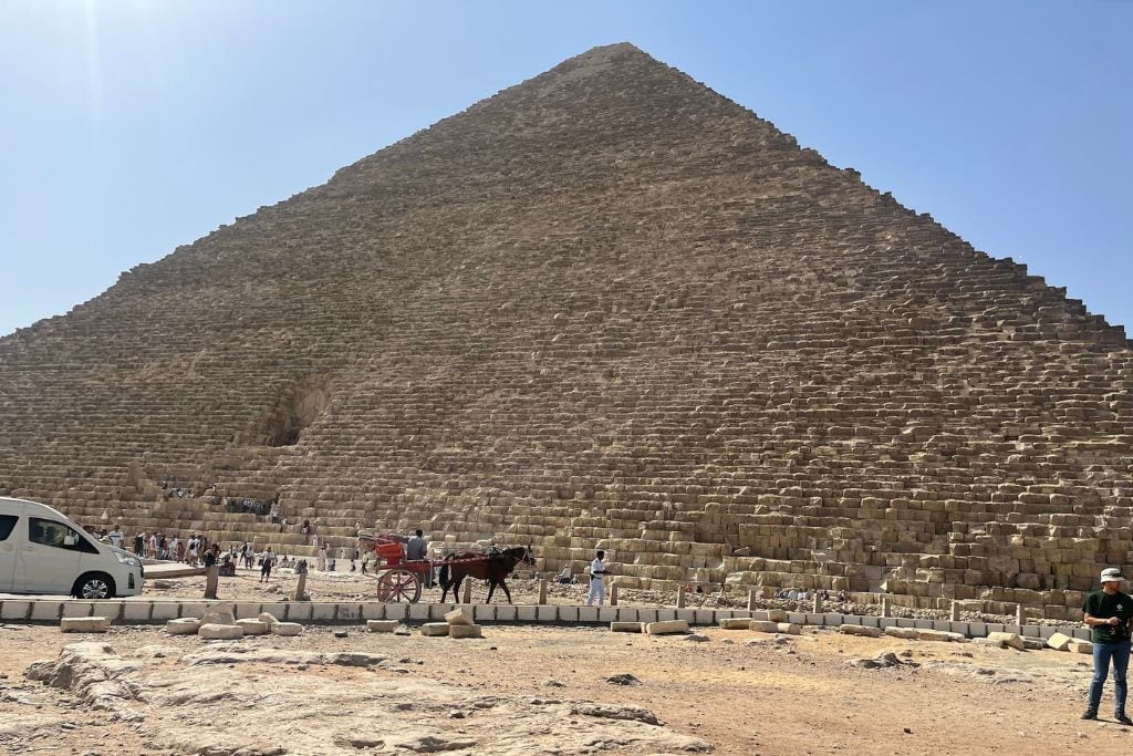 What Do Giza Pyramids Represent Spiritually? The Great Pyramid of Giza looms large, with its immense stone blocks creating a steep, angular facade under a bright sky. Visitors are seen at the base of the pyramid, dwarfed by its size, while a horse-drawn carriage and a parked van are visible nearby. This pyramid, representing the spiritual journey to the afterlife in ancient Egyptian beliefs, stands as a powerful symbol of eternity and the divine connection between the earth and the heavens.