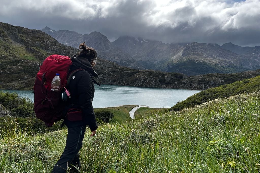 A hiker with a large red backpack stands on a grassy slope overlooking a serene lake surrounded by rugged mountains. The overcast sky and dramatic clouds add a sense of adventure and solitude to the scene, highlighting the remote and wild beauty of the landscape.