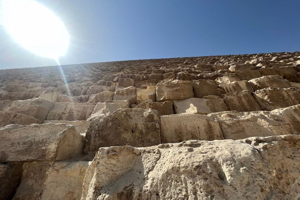What Do Giza Pyramids Represent Spiritually? Close-up view of the massive stone blocks that make up the Great Pyramid of Giza, with the bright sun shining overhead against a clear blue sky. The rough texture and sheer scale of the stones emphasize the monumental effort and spiritual significance behind the construction, symbolizing the pharaoh’s journey to eternity and the connection between earthly life and the divine.