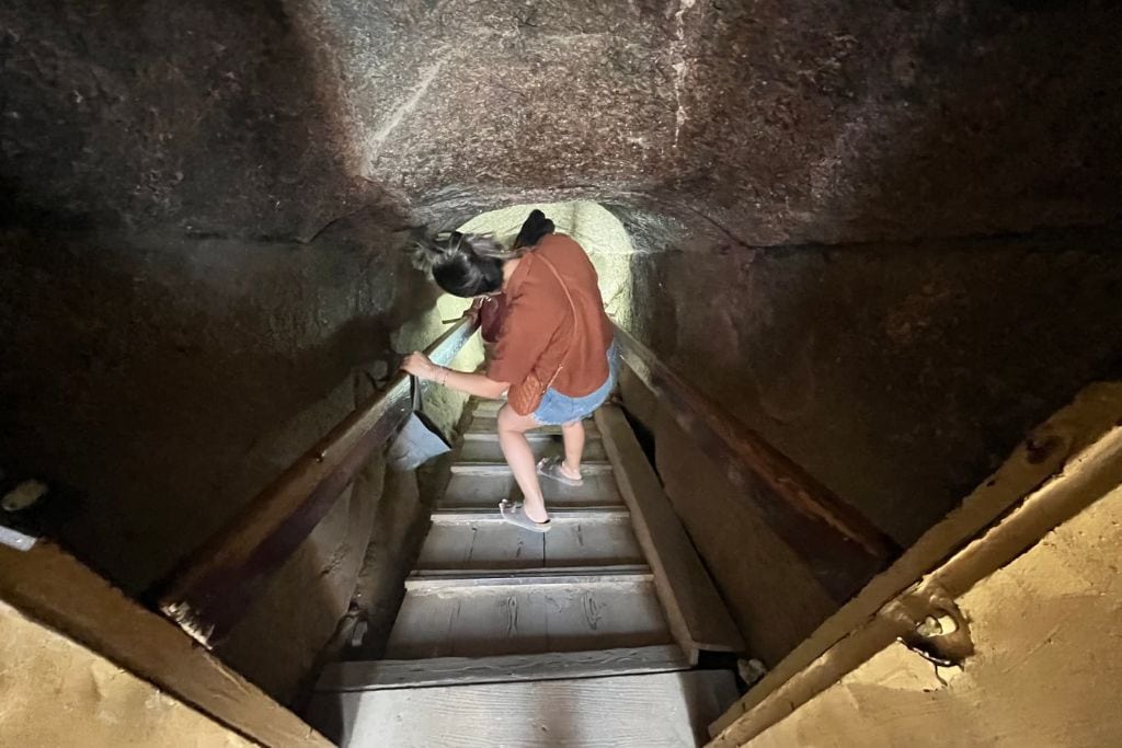 What Do Giza Pyramids Represent Spiritually? A visitor descends a narrow, steep staircase inside one of the pyramids in Egypt, with low ceilings and rough stone walls surrounding the passage. The dimly lit environment and confined space convey the ancient and mysterious atmosphere of the pyramid's interior, reflecting the spiritual significance of these structures as gateways to the afterlife in ancient Egyptian culture.
