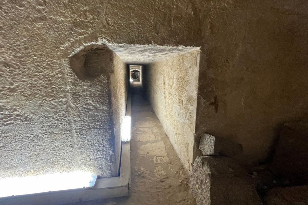 What Do Giza Pyramids Represent Spiritually? A narrow, dimly lit passage inside the Pyramids of Giza, with rough stone walls and a low ceiling. The corridor extends straight ahead, creating a tunnel-like effect, with small lights placed along the floor to guide the way. This enclosed space adds to the mysterious and sacred atmosphere of the pyramid's interior, reflecting its role as a spiritual and ceremonial site in ancient Egyptian culture.