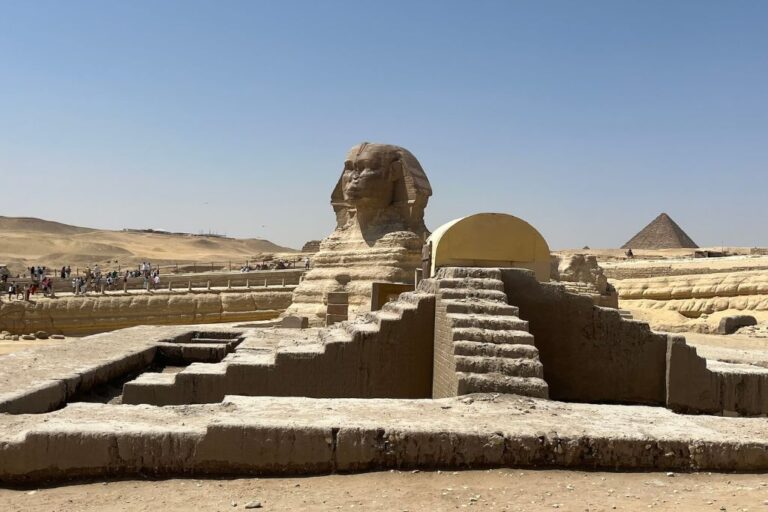The Great Sphinx of Giza stands prominently in the foreground with the Pyramid of Khafre visible in the background. The image captures the iconic monuments under a clear sky with scattered tourists exploring the site. The Giza Pyramids, representing profound spiritual concepts in ancient Egyptian culture, are often seen as symbols of the soul's journey to the afterlife, with the Great Sphinx guarding this sacred space.