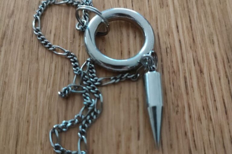 A silver pendulum with a pointed, conical-shaped weight attached to a chain. The pendulum's design features a smooth ring at the top, and it's laid out on a wooden surface.