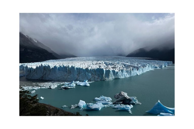 A vast glacier in Patagonia stretches across a turquoise lake, with chunks of ice floating in the water. The dramatic, misty mountains in the background add to the region's rugged beauty, highlighting why Patagonia is worth visiting for its stunning natural landscapes.