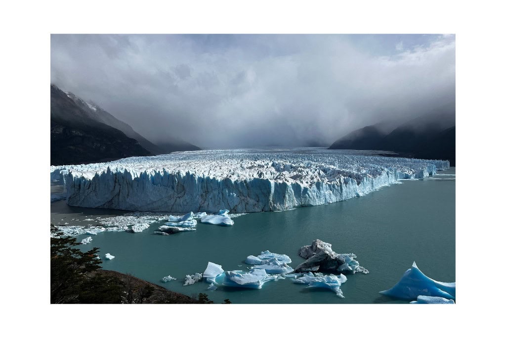 A vast glacier in Patagonia stretches across a turquoise lake, with chunks of ice floating in the water. The dramatic, misty mountains in the background add to the region's rugged beauty, highlighting why Patagonia is worth visiting for its stunning natural landscapes.