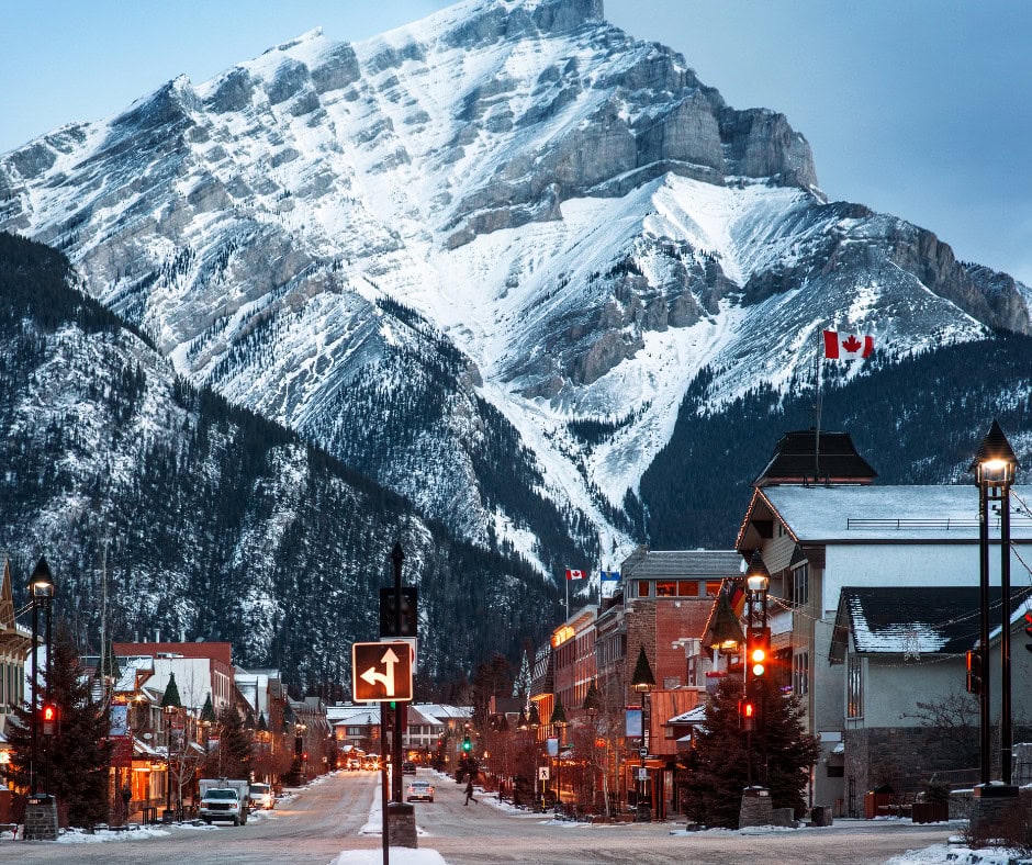 Is Banff Worth Visiting? Explore This Spiritual Place in 2026 A picturesque view of downtown Banff, Canada, with snow-covered buildings lining the street and the towering, snow-capped Cascade Mountain in the background. The Canadian flag flies above the town, creating a cozy, wintery atmosphere under a clear sky.