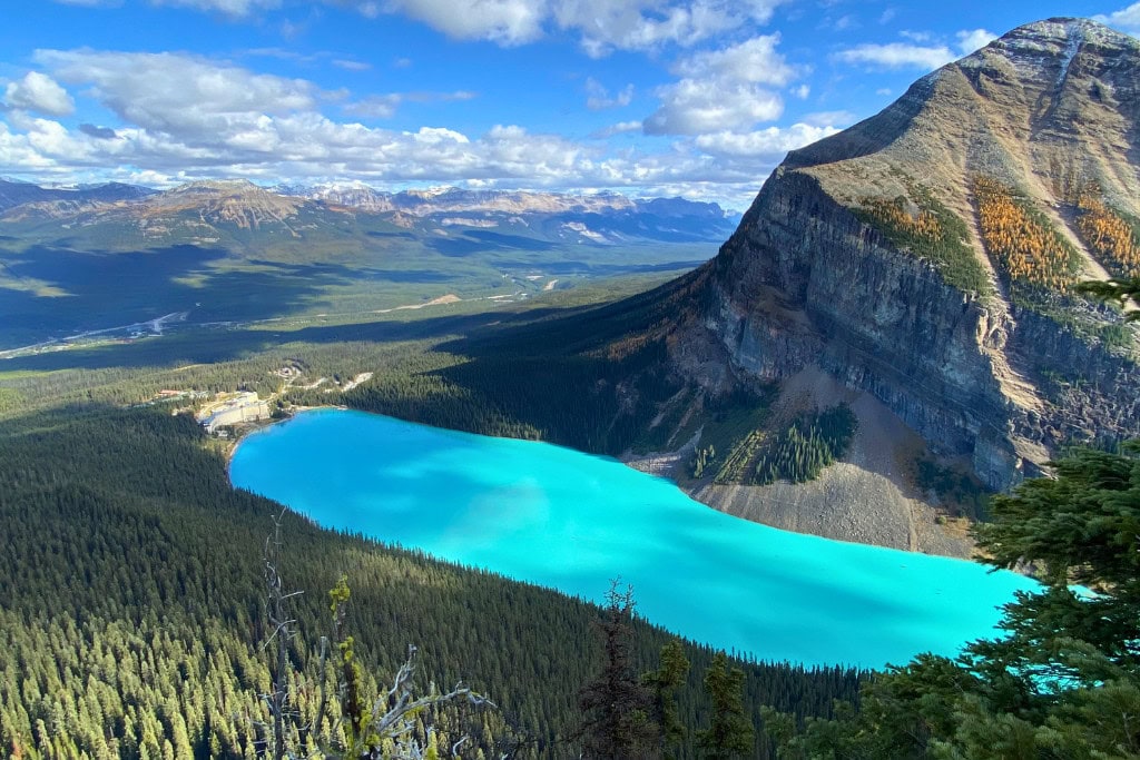 Is Banff Worth Visiting? Explore This Spiritual Place in 2026 A stunning aerial view of Lake Louise in Banff National Park, as seen from the Big Beehive, with its vibrant turquoise waters surrounded by dense forest and towering mountain peaks. The lake's iconic Fairmont Chateau is visible at the shoreline, with expansive mountain ranges extending into the distance under a partly cloudy sky.