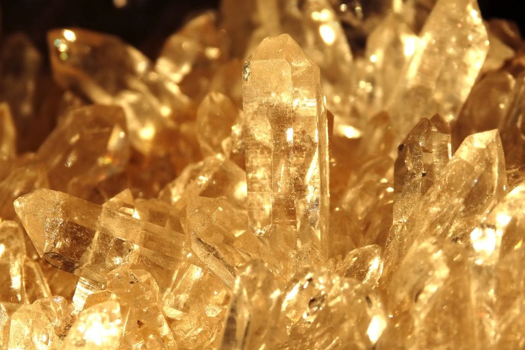 The image shows a close-up of a cluster of golden citrine crystals, glowing with a warm, amber light. The crystals have sharp, pointed tips and are densely packed together, reflecting the light to create a radiant, sparkling effect. The overall scene exudes a sense of warmth and energy due to the rich golden hues.