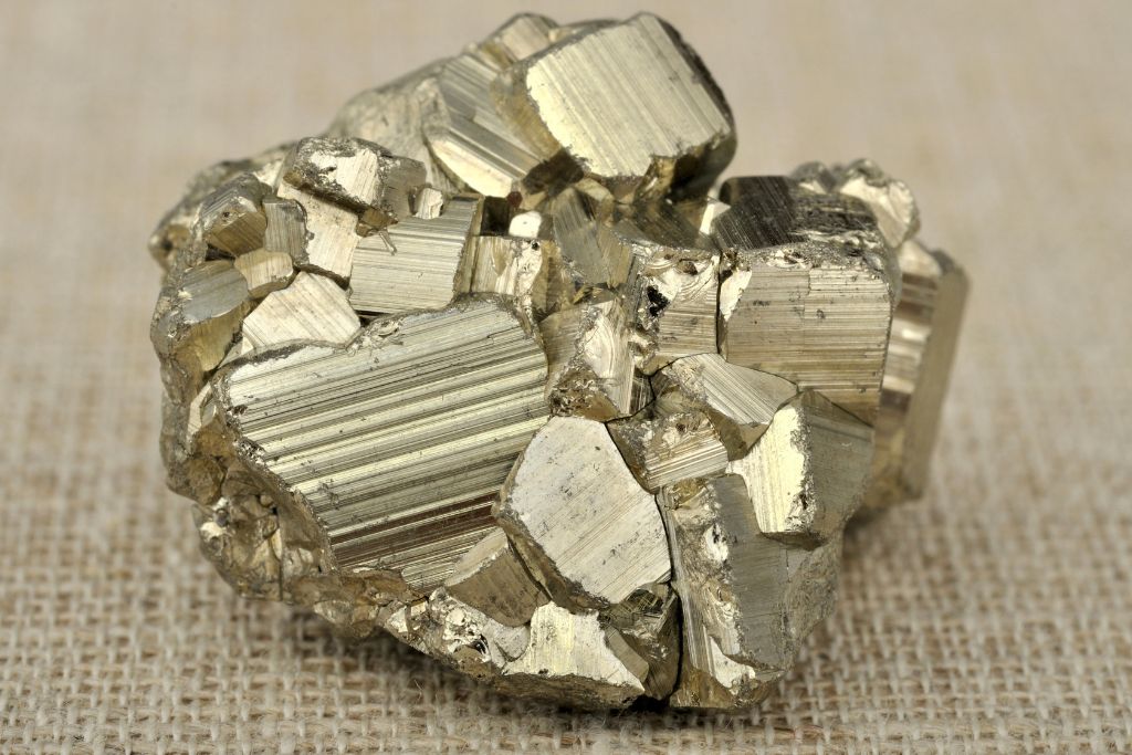 The image shows a cluster of shiny pyrite crystals, also known as "fool's gold," resting on a textured beige surface. The crystals have a metallic, golden sheen with geometric, cubic formations, showcasing striated lines and a reflective surface that catches the light.