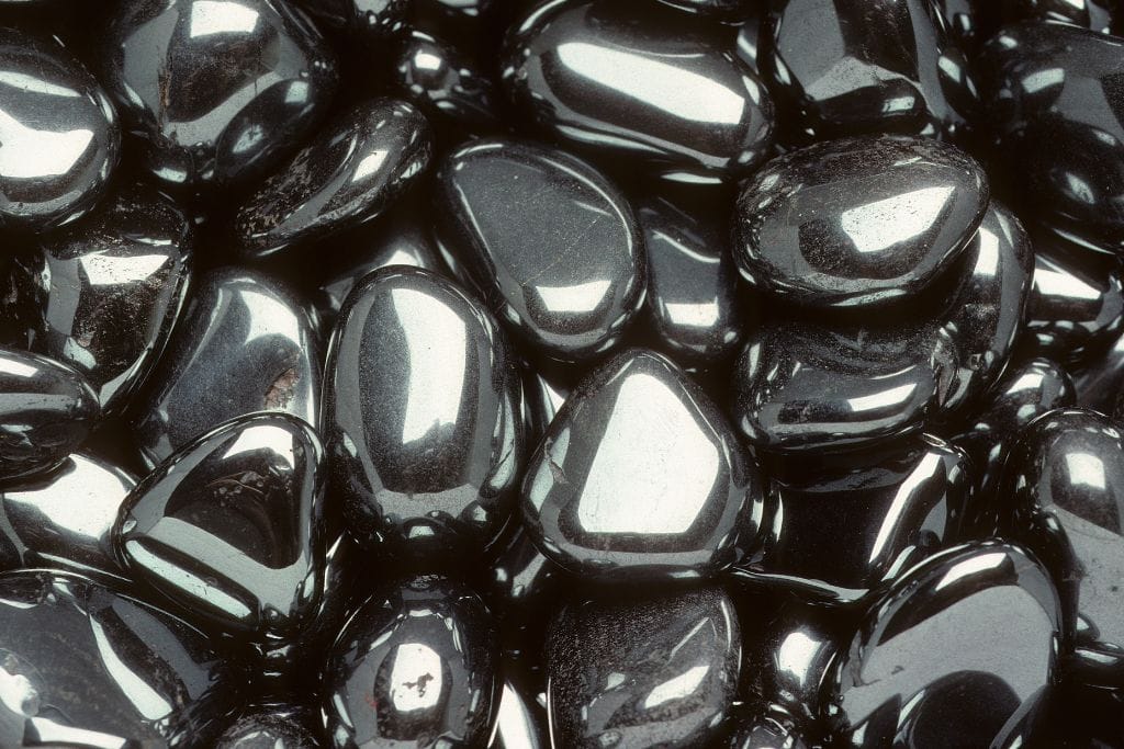 The image displays a close-up of a collection of smooth, polished hematite stones. The stones are metallic black with a glossy, reflective surface, and their shapes vary from rounded to slightly angular. The highly reflective sheen on the stones captures highlights of light, creating a shiny, mirror-like effect across the surfaces.
