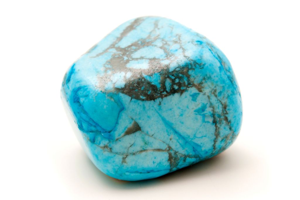 The image shows a polished turquoise stone with vibrant blue hues and intricate veining of black and gray throughout its surface. The stone has a smooth, rounded shape and is placed on a clean, white background, allowing the striking colors and natural patterns to stand out.