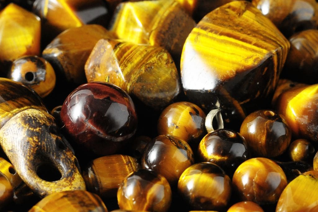 The image shows a collection of polished tiger's eye stones in various shapes and sizes, including beads and rounded forms. The stones exhibit their characteristic golden-brown, honey, and dark brown stripes with a chatoyant, reflective effect that shifts with the light. Among them is a single dark red-brown stone, adding contrast to the overall composition.